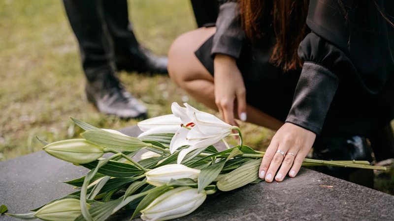 Person kneeling to place a bouquet of white lilies on a gravestone, with another person standing nearby. Scene is set in a cemetery or memorial site.